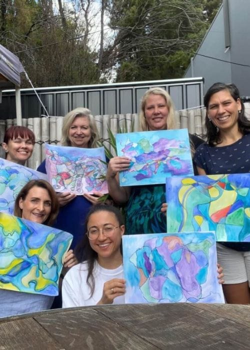 A group of six women smiling and holding up their colorful abstract paintings after an art therapy session at Eltham Wellness Centre. The outdoor setting features a bamboo fence and greenery, creating a relaxing and creative atmosphere.