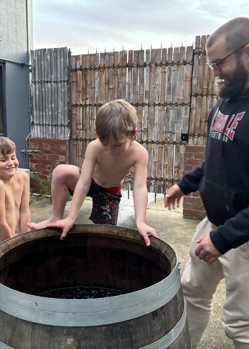 Two young boys and an adult instructor engaging in a cold exposure session at Eltham Wellness Centre. One child prepares to step into a rustic wooden ice bath, while the instructor smiles and guides them through the experience in an outdoor setting.