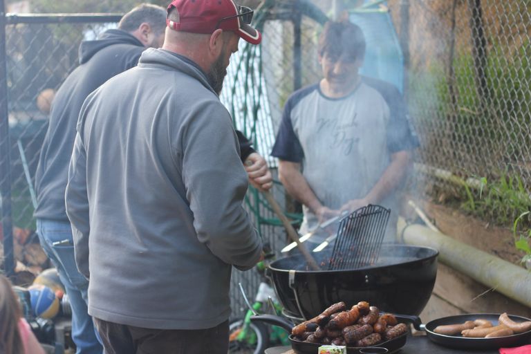 A group of people enjoying a community BBQ at Eltham Wellness Centre. Smoke rises from the grill as participants cook sausages and chat, creating a friendly and social outdoor gathering.