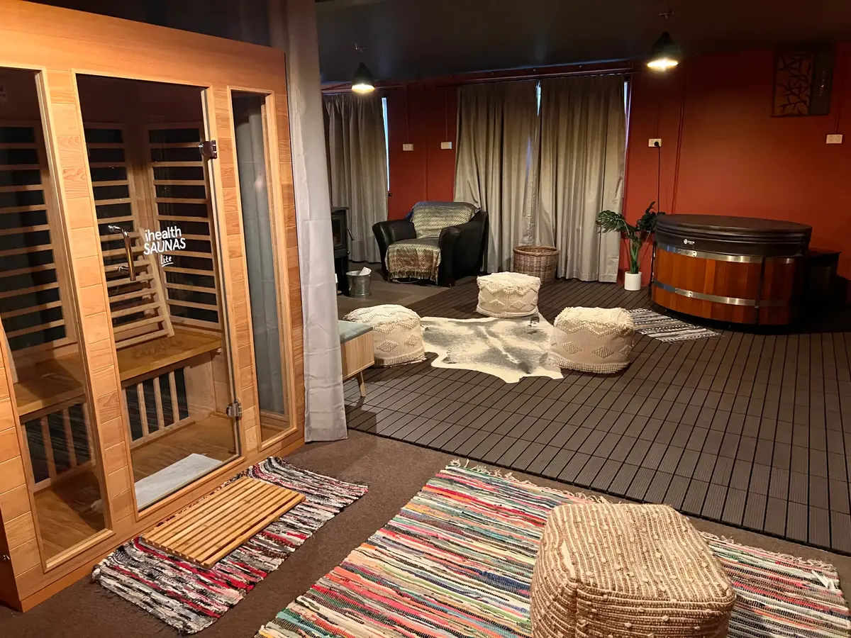 A cozy contrast therapy room at Eltham Wellness Centre, featuring an infrared sauna, a wooden hot tub, and a relaxing lounge area. The space is designed with warm lighting, earthy tones, and soft furnishings, including woven ottomans, a cowhide rug, and comfortable seating. The serene ambiance provides an ideal setting for heat and cold therapy, promoting relaxation and recovery.