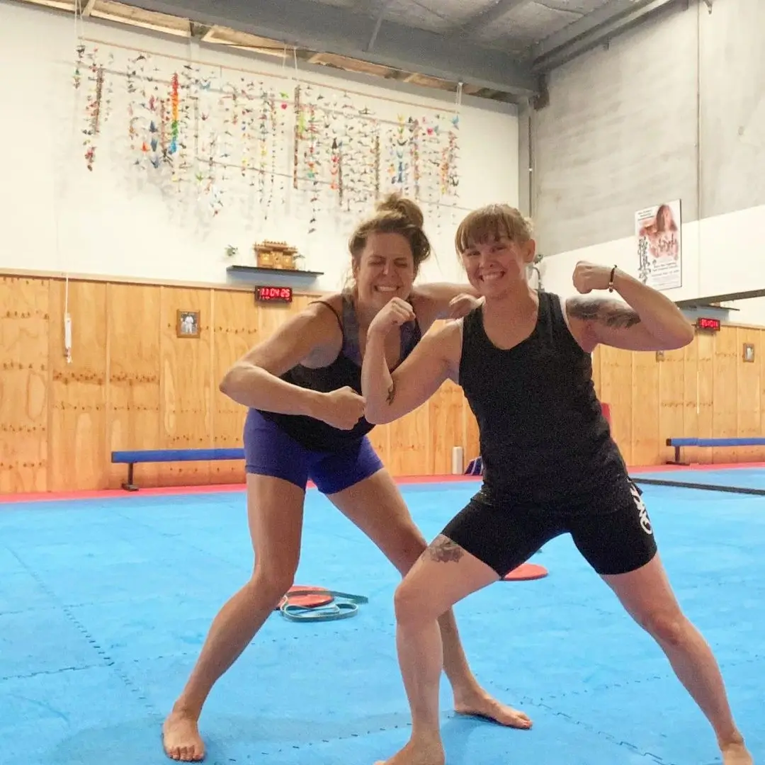 Two women demonstrating corrective exercises as part of a myotherapy session at Eltham Wellness Centre. They are standing on blue mats in a spacious training area, flexing and engaging in movement-based therapy. The background features wooden walls, colorful hanging decorations, and training equipment, creating a dynamic and supportive environment for injury prevention and rehabilitation.