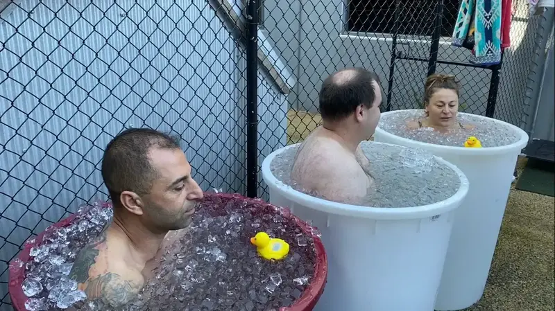 Three people sitting in large ice baths at Eltham Wellness Centre, immersed in freezing water with ice floating on the surface. Each person is focused and calm, practicing breath control to manage the cold exposure. A small rubber duck floats in each tub, adding a lighthearted touch to the scene. The setting is outdoors, enclosed by a black fence, creating a communal yet private space for cold therapy and recovery.