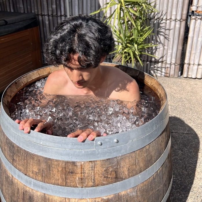 A young man with dark curly hair sits in a rustic wooden barrel filled with ice during an outdoor ice bath therapy session. His hands grip the edge of the barrel as he focuses on his breath. The setting includes a bamboo fence, green plants, and a warm, sunny atmosphere, enhancing the contrast between the cold water and the surrounding environment.