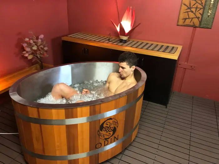 A man sits in a large wooden ice bath filled with ice cubes at Eltham Wellness Centre, focusing on his breath as he undergoes cold therapy. The room has a warm, inviting ambiance with soft lighting, decorative plants, and a calming red-toned interior. The contrast between the icy water and the cozy surroundings highlights the benefits of cold immersion for recovery and resilience.