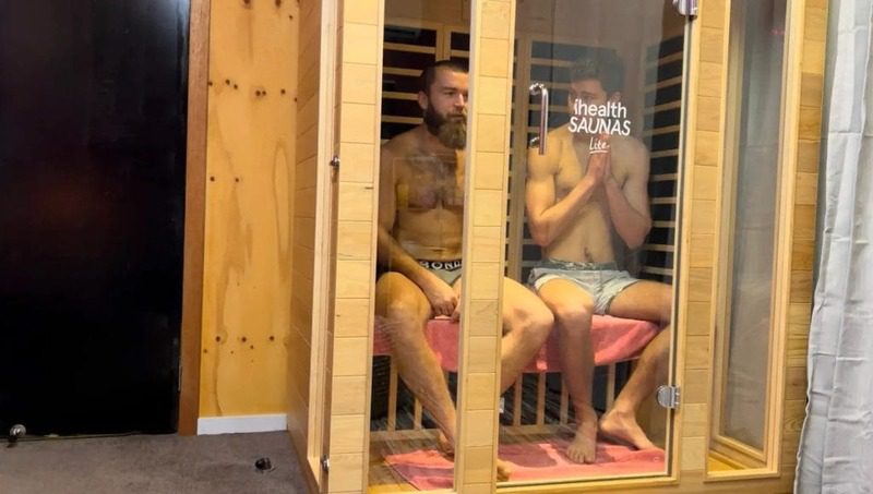 Two men sitting inside an infrared sauna at Eltham Wellness Centre, engaging in heat therapy. One man sits calmly with a focused expression, while the other has his hands near his face, possibly adjusting to the warmth. The wooden sauna cabin has glass doors and infrared heating panels, creating a cozy and therapeutic environment for relaxation and recovery.