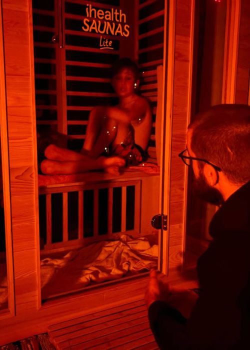 A warmly lit infrared sauna session at Eltham Wellness Centre. A person sits inside the sauna, bathed in red light, while an instructor or guide observes from outside, creating an intimate and relaxing therapy experience