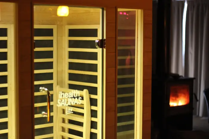 A warmly lit infrared sauna at Eltham Wellness Centre, showcasing its sleek wooden design and glass door. The sauna emits a gentle glow, creating a relaxing atmosphere. In the background, a wood-burning fireplace adds to the cozy ambiance, enhancing the wellness experience.