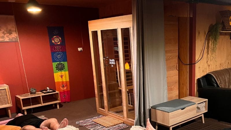 A cozy wellness space at Eltham Wellness Centre featuring a wooden infrared sauna, a comfortable lounge area, and chakra wall art. The room has warm lighting, soft furnishings, and a relaxing ambiance. In the foreground, individuals are lying down, possibly engaging in a breathwork or meditation session, enhancing the holistic wellness experience.