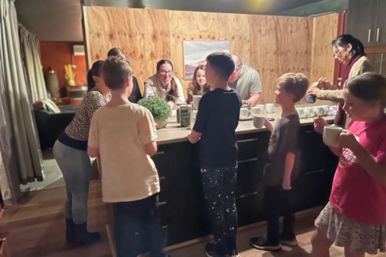 A group of adults and children gathered around a kitchen counter at Eltham Wellness Centre, enjoying tea and conversation. The warm lighting and wooden interior create a cozy and inviting atmosphere.