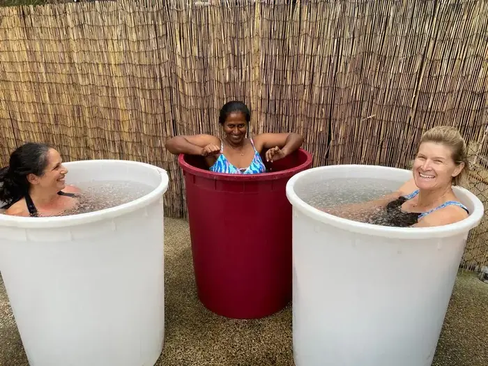 Three women smiling and enjoying an outdoor ice bath session at Eltham Wellness Centre. They are each immersed in large plastic tubs filled with cold water, with a bamboo fence in the background providing a tranquil and private setting. The women appear to be having fun, embracing the benefits of cold exposure for wellness and recovery.