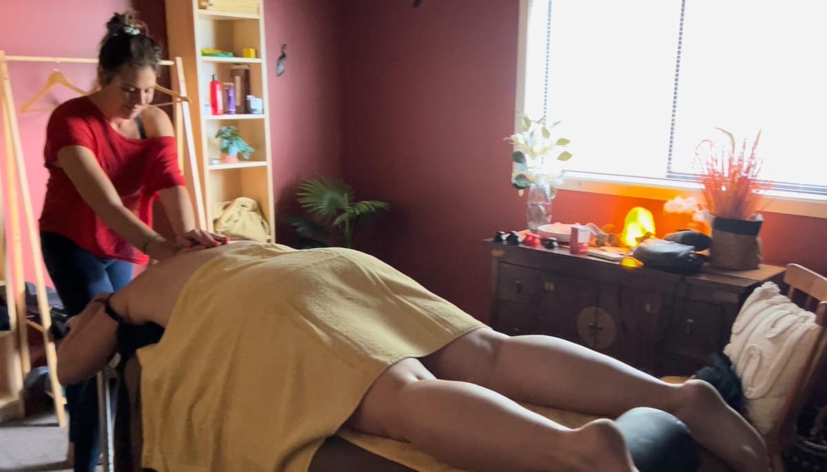A professional massage therapist providing a remedial massage in a cozy, well-lit treatment room at Eltham Wellness Centre. The client is lying face down on a massage table, covered with a towel, while the therapist applies pressure to release muscle tension. The peaceful ambiance includes soft lighting, plants, and a Himalayan salt lamp.