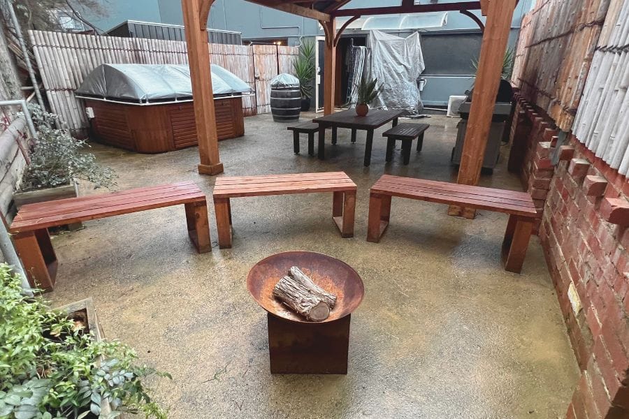 Outdoor wellness area at Eltham Wellness Centre featuring wooden benches, a central fire pit, covered seating, and an infrared sauna, designed for relaxation and recovery.