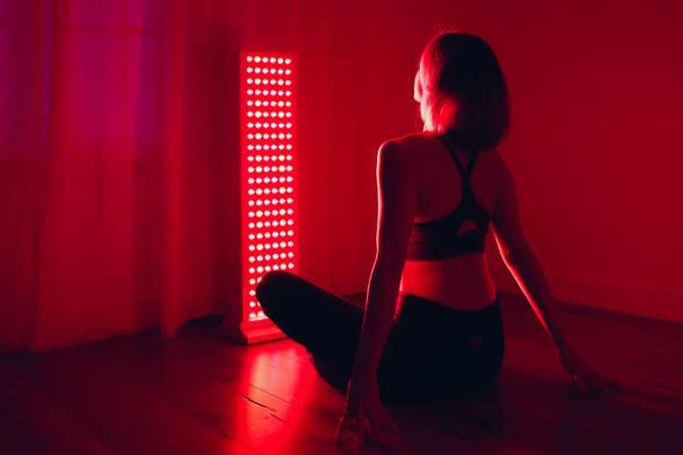 Red Light Therapy for Recovery & Relaxation – Eltham Wellness Centre A woman sits in front of a full-body red light therapy panel in a dark, peaceful room, promoting muscle recovery, skin health, and relaxation.