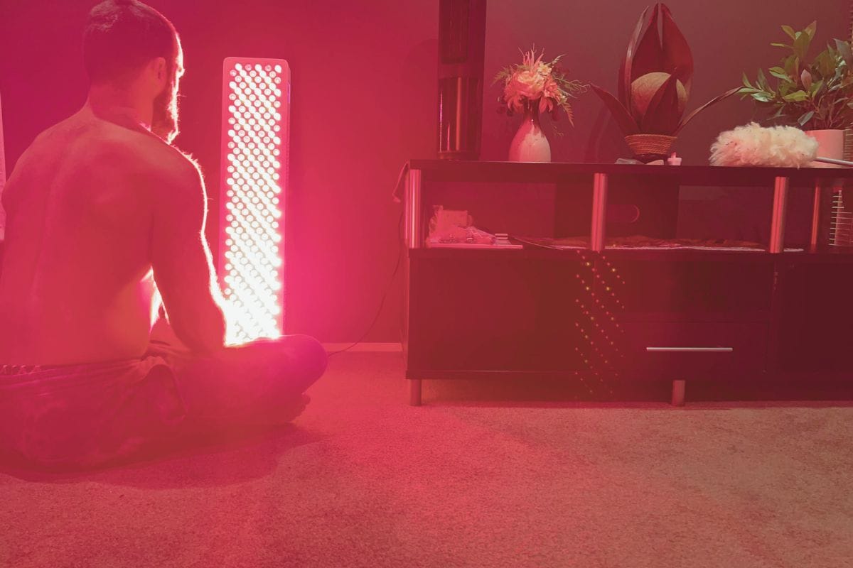 A man sits cross-legged in front of a full-body red light therapy panel at Eltham Wellness Centre, surrounded by soft lighting and calming decor.