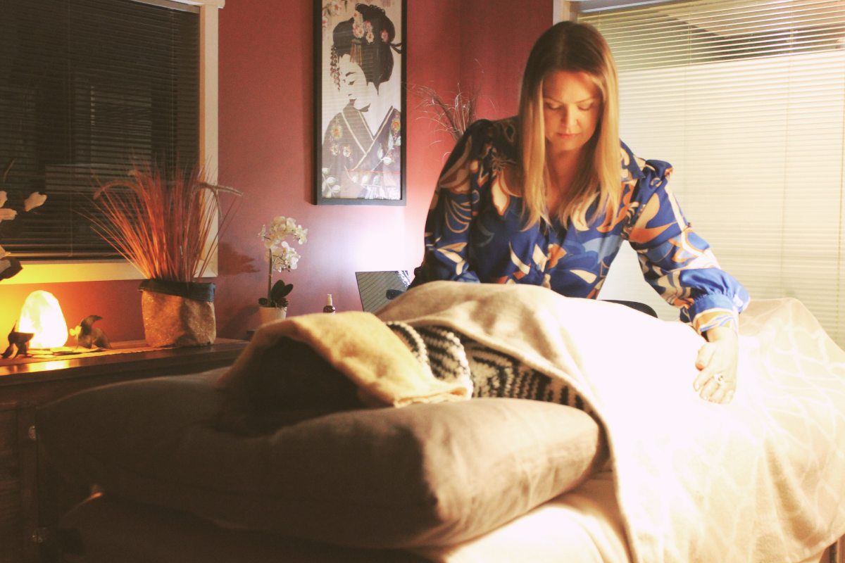 A Reiki practitioner gently placing hands over a client covered with a blanket in a softly lit wellness room with warm decor and a framed artwork on the wall.